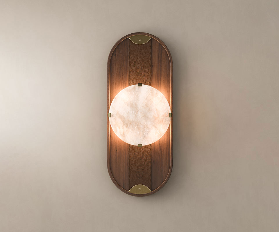Cavendish Wall Lamp, front view, with the light turned on. Crafted from smoked walnut wood and alabaster, framed in a gentle oval silhouette with aged brushed brass accents, showcasing the lamp’s refined materials and elegant design set against a beige wall.