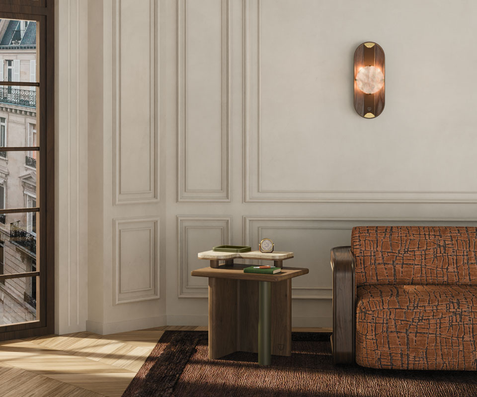 Maurier side table with a cylindrical leather-wrapped leg, wooden frame, and smooth travertine tabletop stands beside a window, paired with the Kimberley sofa upholstered in Velours Rocca by Nobilis, featuring a smoked walnut structure and rattan details. Above, the Cavendish wall lamp in smoked walnut, alabaster, leather, and aged brushed brass casts a warm glow, enriching the serene, refined ambience. This pieces are brand new arrivals in the collection.