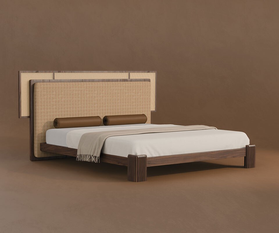 Forbes bed rattan bedroom furniture. Forbes bed in a 45º front view, featuring a fabric and rattan headboard, cylindrical leather bolster pillows, and a walnut wood frame, set against a brown background.
