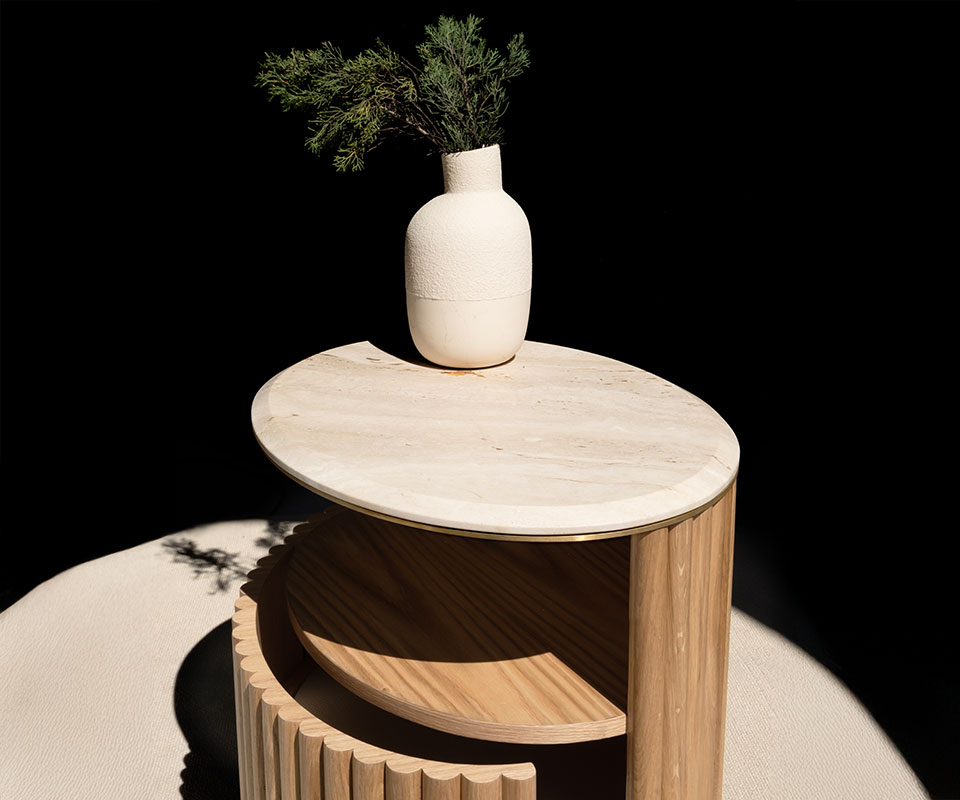 Herbert Bedside Table in natural oak with a detailed travertine tabletop and brushed brass accents, positioned at a 45-degree angle. A white ceramic vase with a plant sits on top. Strong shadows highlight the fluted wood texture, and the door is slightly open.