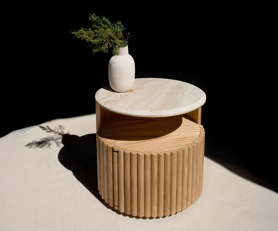 Herbert Bedside Table in natural oak with a travertine tabletop and brushed brass details, positioned at a 45-degree angle. A white ceramic vase with a plant sits on top. Strong shadows enhance the fluted wood texture.