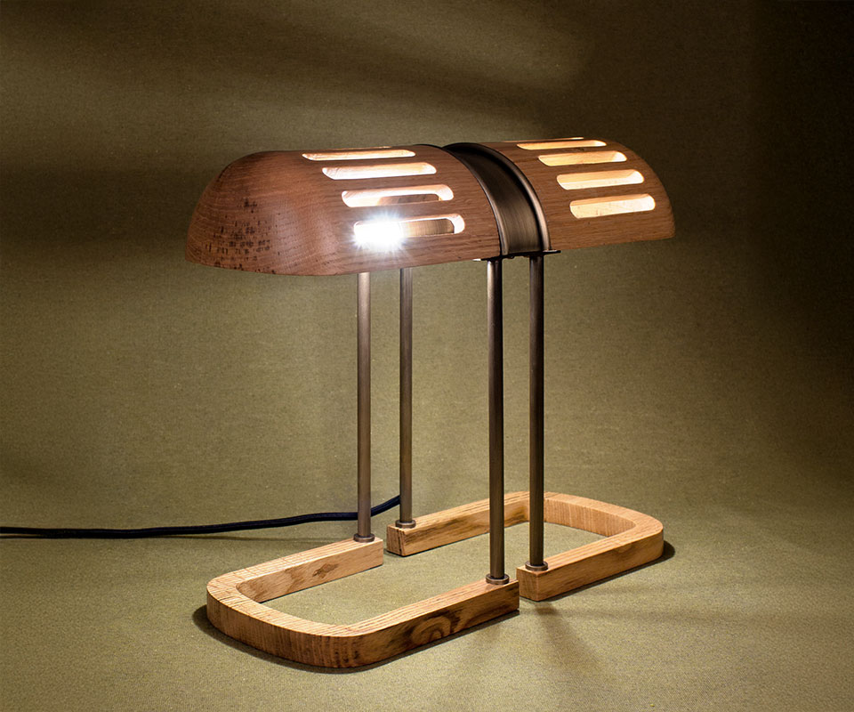 Turing Table Lamp crafted in natural oak wood with aged brass accents, lit with the shade casting beautiful shadows against an olive green background.